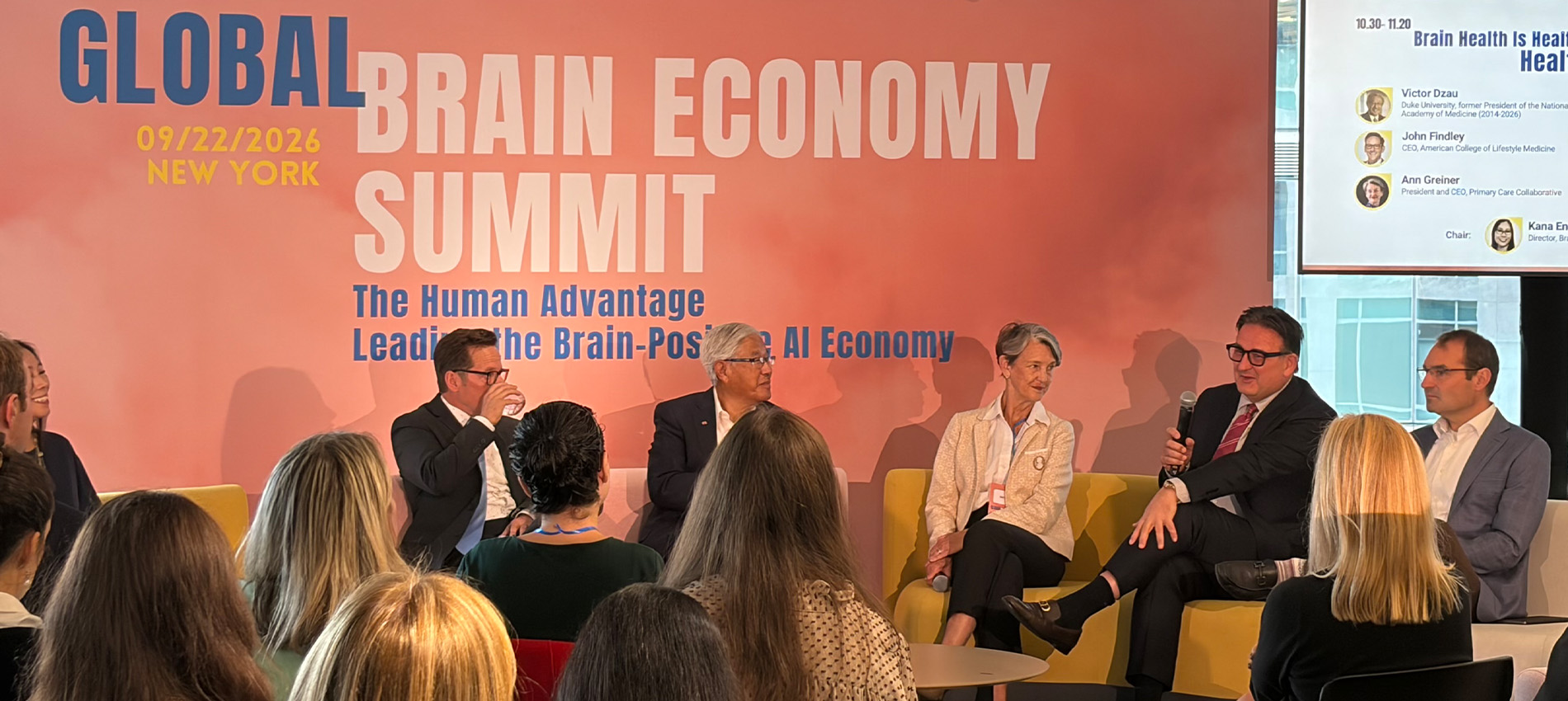 Five people are shown seated on stage before an audience and a presentation screen displayed to the side during a panel discussion at the Global Brain Economy Summit in New York.