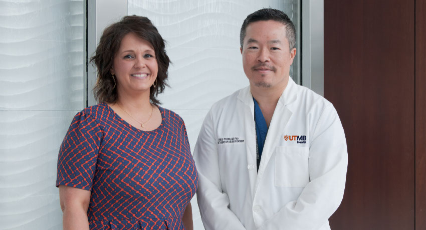 Two people standing side by side indoors; one wears a patterned short-sleeve dress, and the other wears a white lab coat with a UTMB logo over blue scrubs, positioned in front of a light textured wall and window.