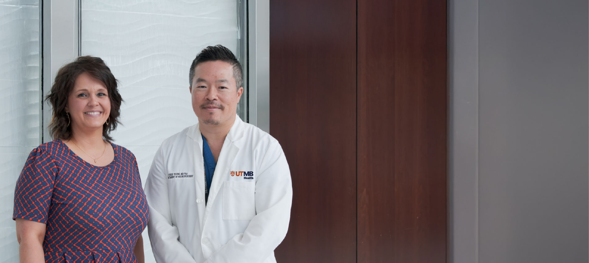 Two people standing side by side indoors; one wears a patterned short-sleeve dress, and the other wears a white lab coat with a UTMB logo over blue scrubs, positioned in front of a light textured wall and window.