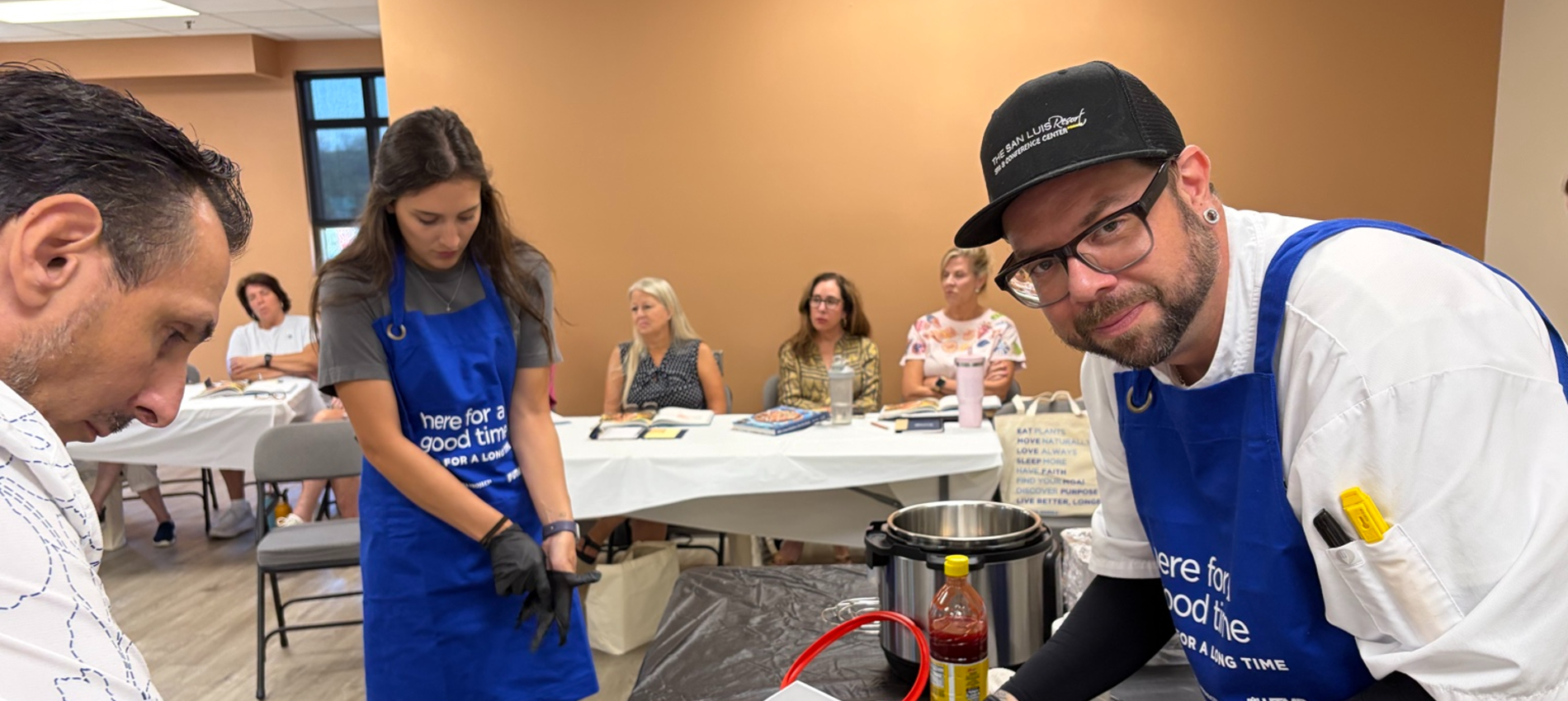 Three people wearing blue aprons stand around a table with cooking equipment and ingredients, while several people seated at tables watch in the background.