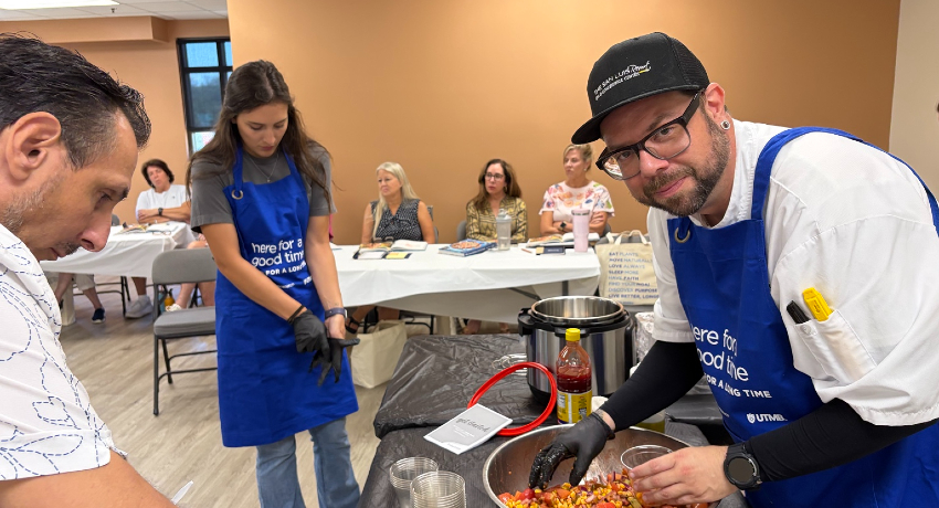 Three people wearing blue aprons stand around a table with cooking equipment and ingredients, while several people seated at tables watch in the background.