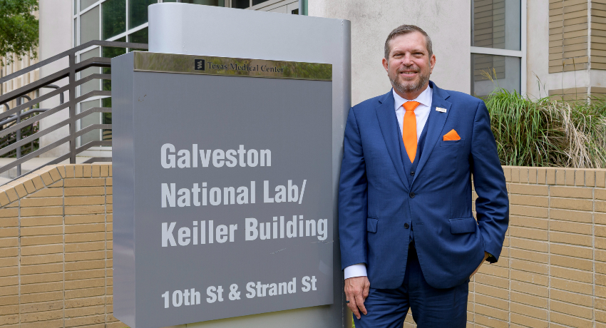 Sign reading ‘Galveston National Lab/Keiller Building, 10th St & Strand St’ stands outside a campus building, with a person in a blue suit standing beside it.