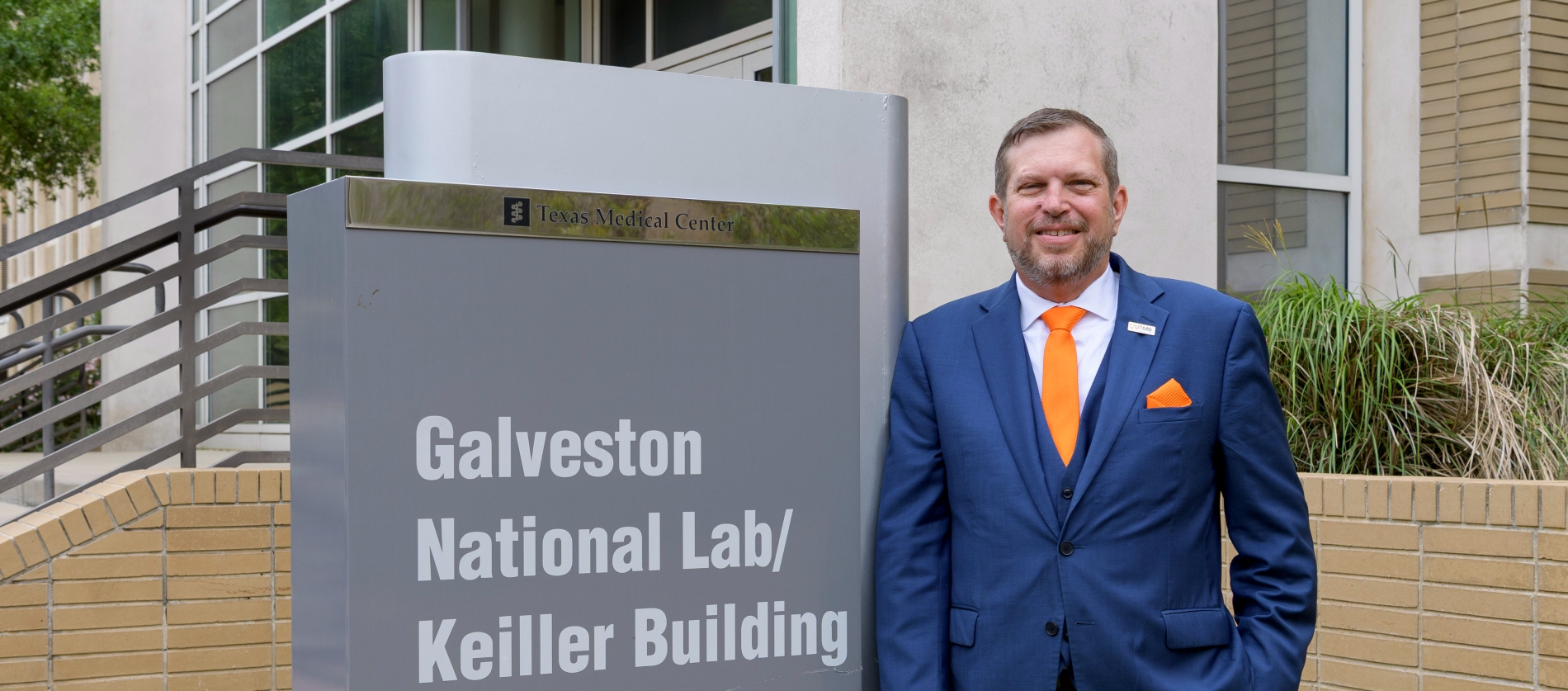 Sign reading ‘Galveston National Lab/Keiller Building, 10th St & Strand St’ stands outside a campus building, with a person in a blue suit standing beside it