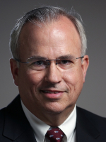 ormal head-and-shoulders portrait of a person wearing a dark suit jacket, white dress shirt, and patterned tie against a neutral gray background.
