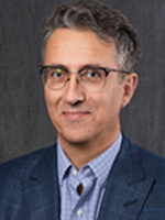 Head-and-shoulders studio portrait of a person wearing a blue blazer over a light blue button-down shirt, photographed against a textured dark gray background.