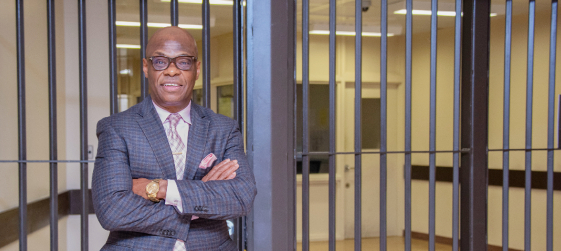 A man in a suit and glasses stands with arms crossed in front of a metal-barred doorway inside a building.