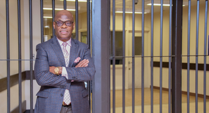 A man in a suit and glasses stands with arms crossed in front of a metal-barred doorway inside a building.