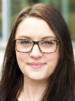  In a head-and-shoulders photo, a woman with long brown hair and glasses is shown against a blurred outdoor background. 