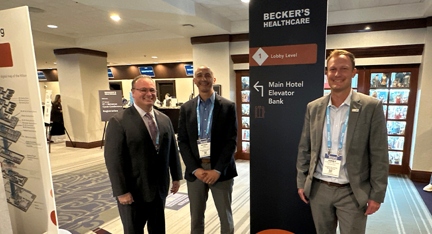 Three conference attendees wearing business attire stand beside a Becker’s Healthcare directional sign in a hotel conference lobby. Registration and conference signage are visible in the background.