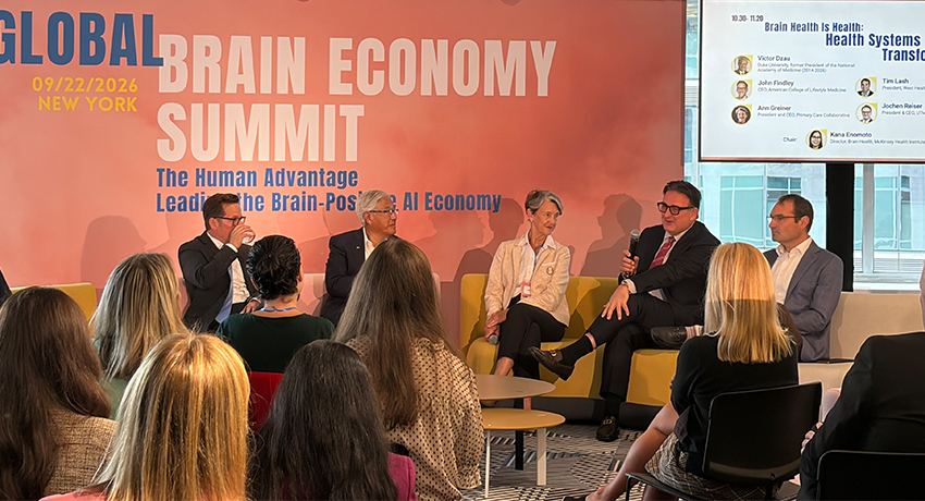 Five people are shown seated on stage before an audience and a presentation screen displayed to the side during a panel discussion at the Global Brain Economy Summit in New York.