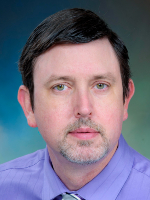 A man is shown in a head-and-shoulders portrait wearing a light purple shirt and striped tie, facing the camera against a blue-green gradient background. 

.