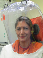 Research staff member wearing an orange shirt inside a clear, sealed positive-pressure hood with external air hose connection in a laboratory setting..
