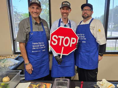 Three people wearing blue UTMB aprons stand behind a table and hold a red octagonal STOP sign. Cooking supplies and food preparation materials are visible on the table.