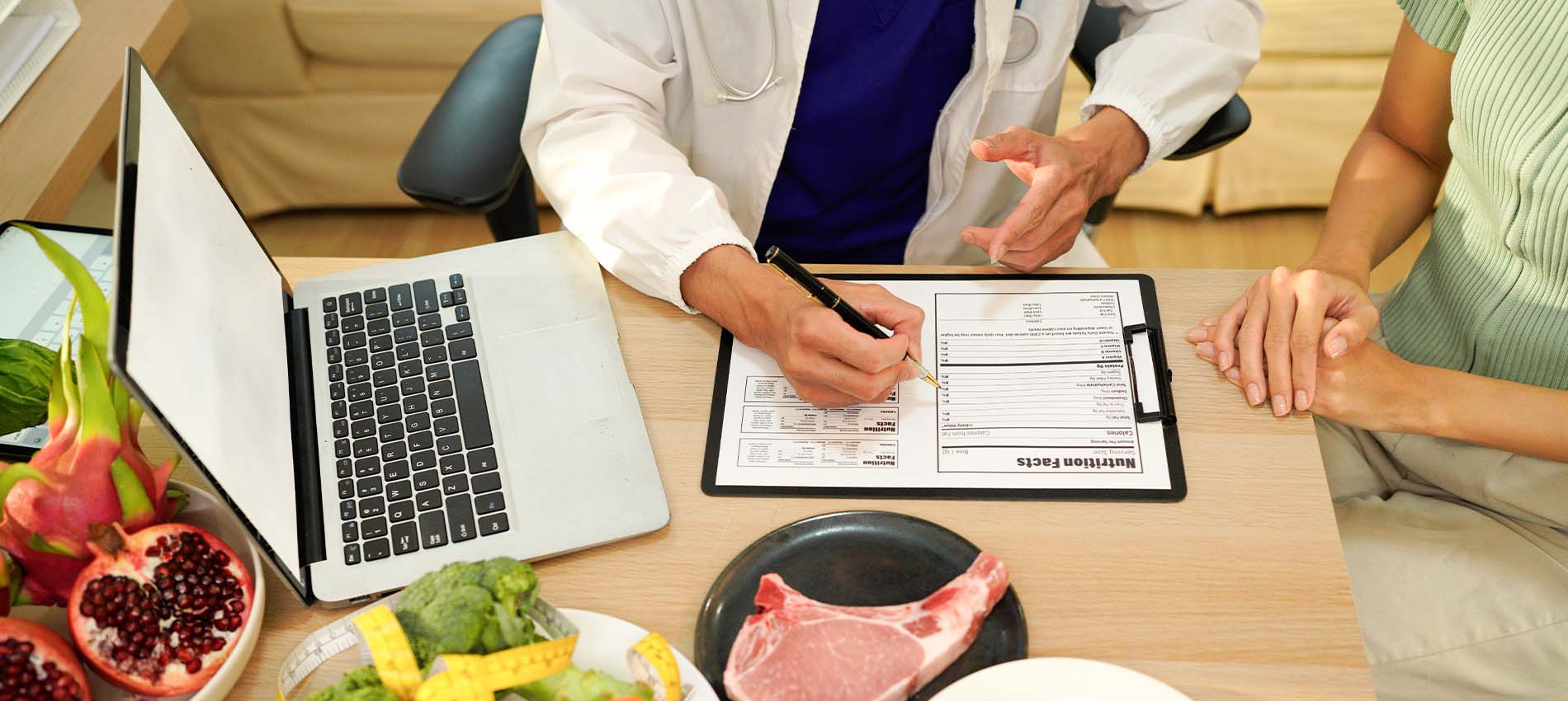 Doctor in a white coat reviews a nutrition plan on a clipboard with a patient at a desk; laptop open beside them, and various foods—fresh vegetables, pomegranate, salmon, and a raw pork chop—are arranged on the table, illustrating a dietary consultation.