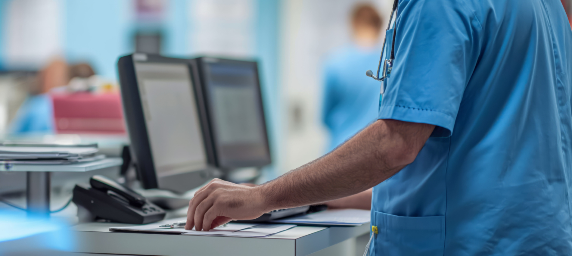 A healthcare worker in blue scrubs is shown reviewing paperwork at a workstation with computer monitors. A patient care area is visible in the background.