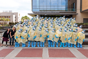 Large group wearing yellow protective gowns, face shields, masks, and blue gloves stand in rows outside a building with signage reading “Health Education Center,” all leaning to one side. 