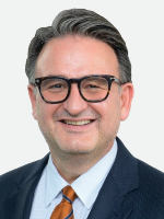 Dr. Jochen Reiser Professional head-and-shoulders photo shows a man with short dark hair and glasses wearing a dark blue suit and orange tie with a light blue shirt.