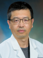  In a head-and-shoulders professional headshot, a man with glasses and short dark hair is wearing a white lab coat over a green shirt 