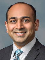  Professional head-and-shoulders photo shows a man with dark hair wearing a dark grey suit, white shirt and blue tie against a gray background. 