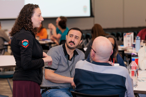 Instructor standing beside a table speaking to a small group of seated participants in a classroom setting, with water bottles and materials on the table. A projection screen and additional attendees are visible in the background.