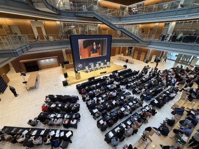 Audience seated in rows in a large multi-level atrium, facing a stage with a panel of speakers and a large screen displaying a presenter at a podium. Upper balconies and walkways overlook the event space.