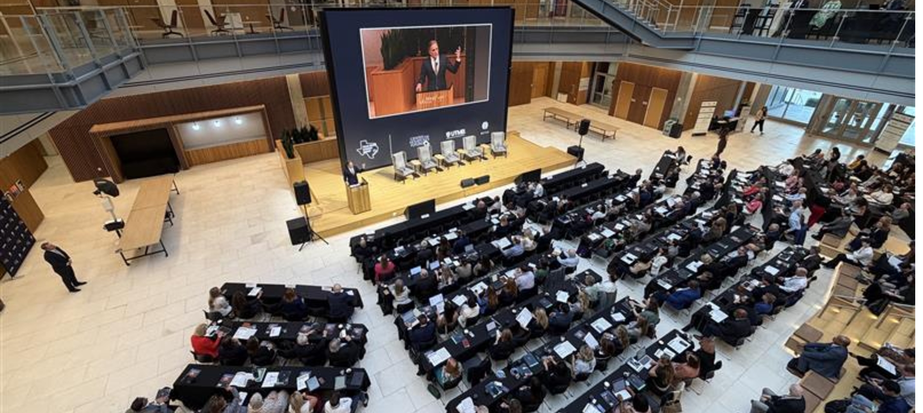 Wide view of an indoor atrium with tiered seating where a large audience sits at tables facing a stage with a podium and a large screen displaying a speaker. Rows of chairs, lighting equipment, and a panel setup are visible on the stage.