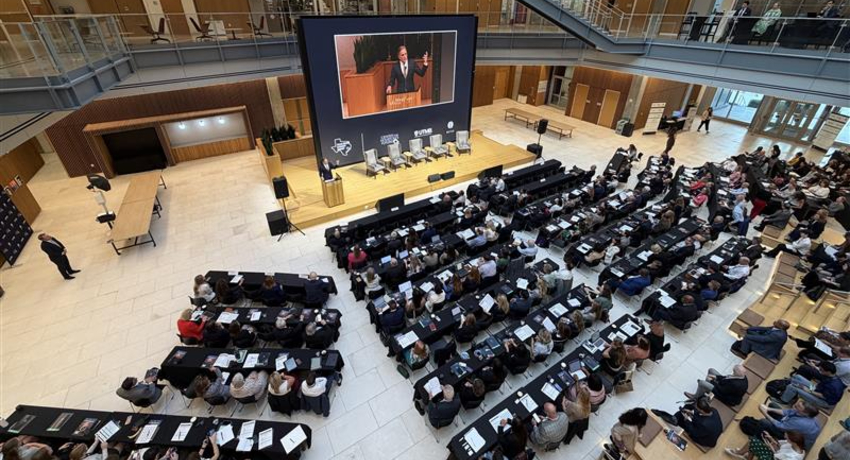 Wide view of an indoor atrium with tiered seating where a large audience sits at tables facing a stage with a podium and a large screen displaying a speaker. Rows of chairs, lighting equipment, and a panel setup are visible on the stage.
