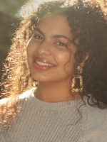 A woman with curly hair is shown in a head-and-shoulders portrait wearing a light-colored sweater, facing the camera outdoors with sunlight filtering through the background.