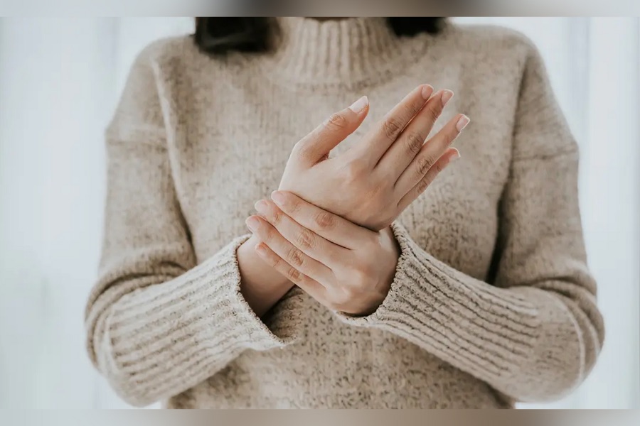 Person wearing a light-colored sweater gently holding one wrist with the opposite hand, focusing on the hands and wrists, suggesting hand or wrist discomfort