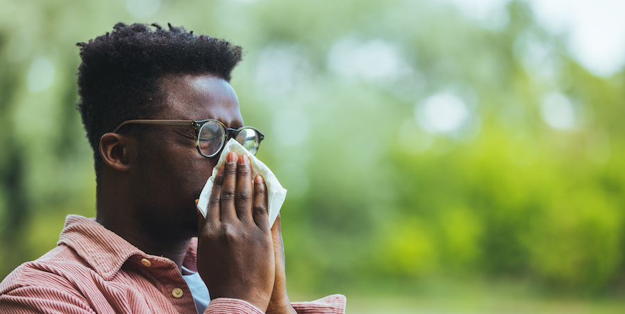 Person wearing glasses and a long-sleeve shirt holding a tissue over their nose outdoors, with green foliage in the background, suggesting sneezing or nasal irritation during allergy season