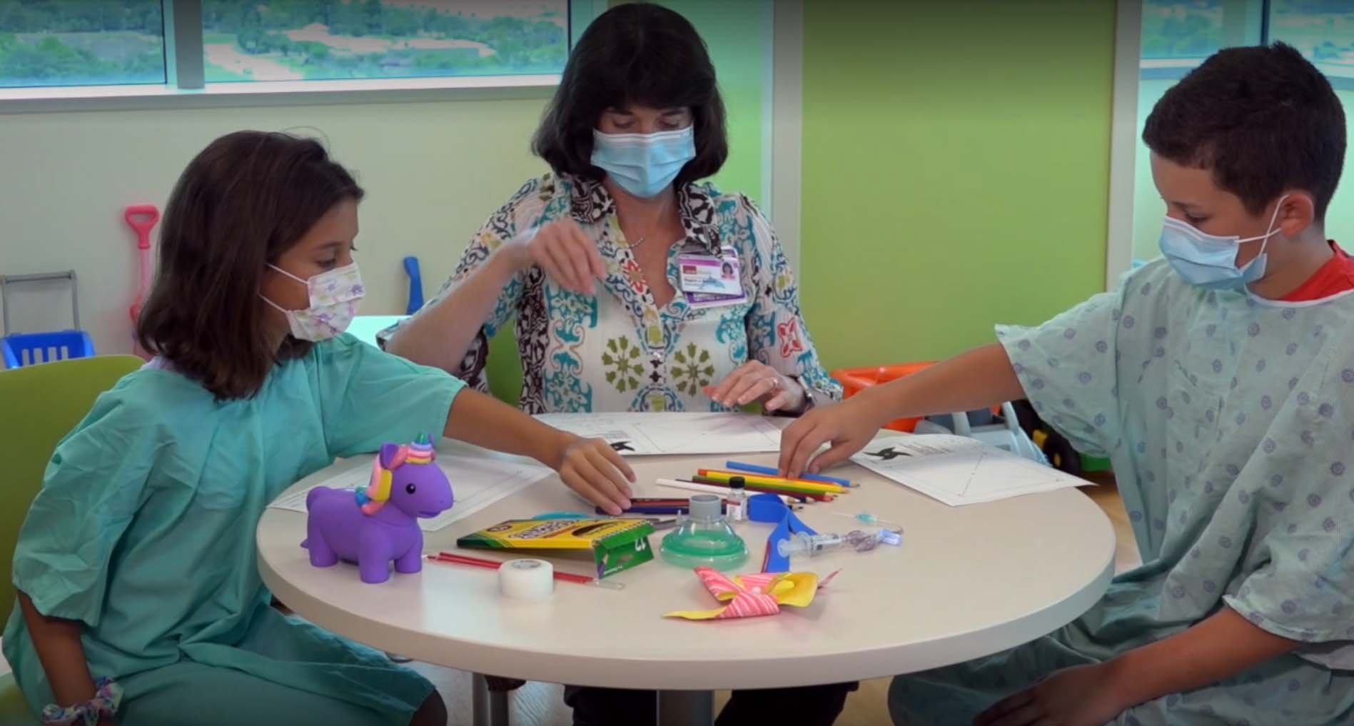 A certified child life specialist sits at a table with two young patients in hospital gowns and masks, guiding them through drawing and play activities to help reduce anxiety during their hospital stay