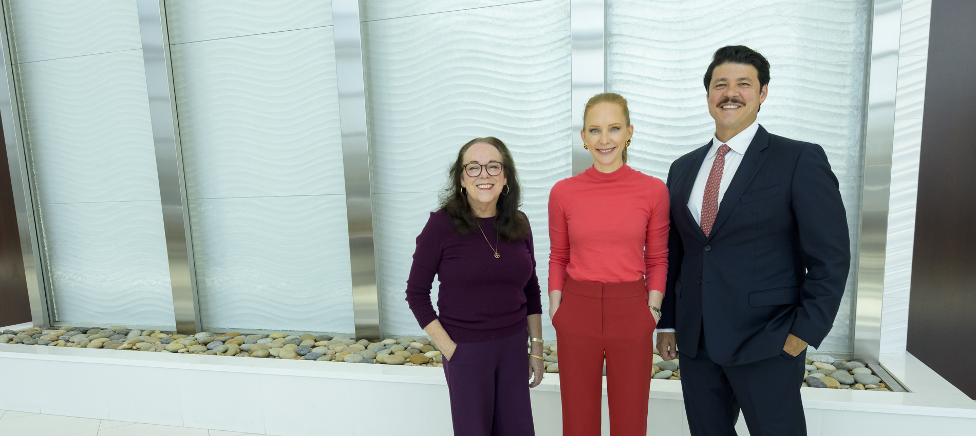 Two women and man are shown standing in front of a white textured wall, with decorative stones visible at the base of the wall. One person wears purple, one wears a bright coral top and matching pants, and one wears a navy suit with a red tie.