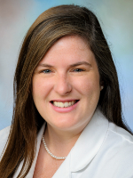 A woman wearing a white coat and patterned top against a blue-green studio background is shown in a head-and-shoulders portrait.