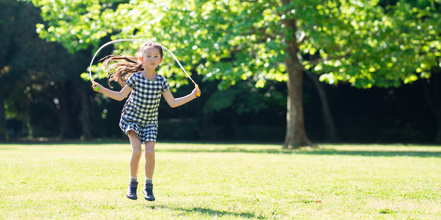 Young girl jumping rope on a grassy field in a park, with trees and sunlight in the background