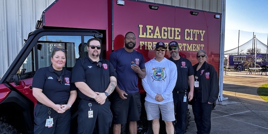 Group of UTMB team members standing with League City first responders in front of a League City Fire Department vehicle during a community youth baseball event