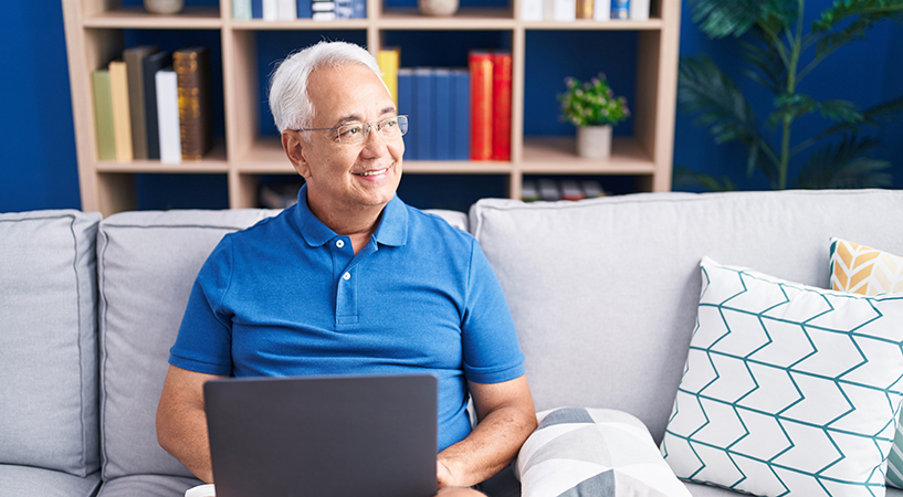Person sitting on a couch at home using a laptop, with a bookshelf and decorative pillows in the background