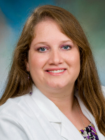 A woman wearing a white coat and patterned top against a blue-green studio background is shown in a head-and-shoulders portrait.