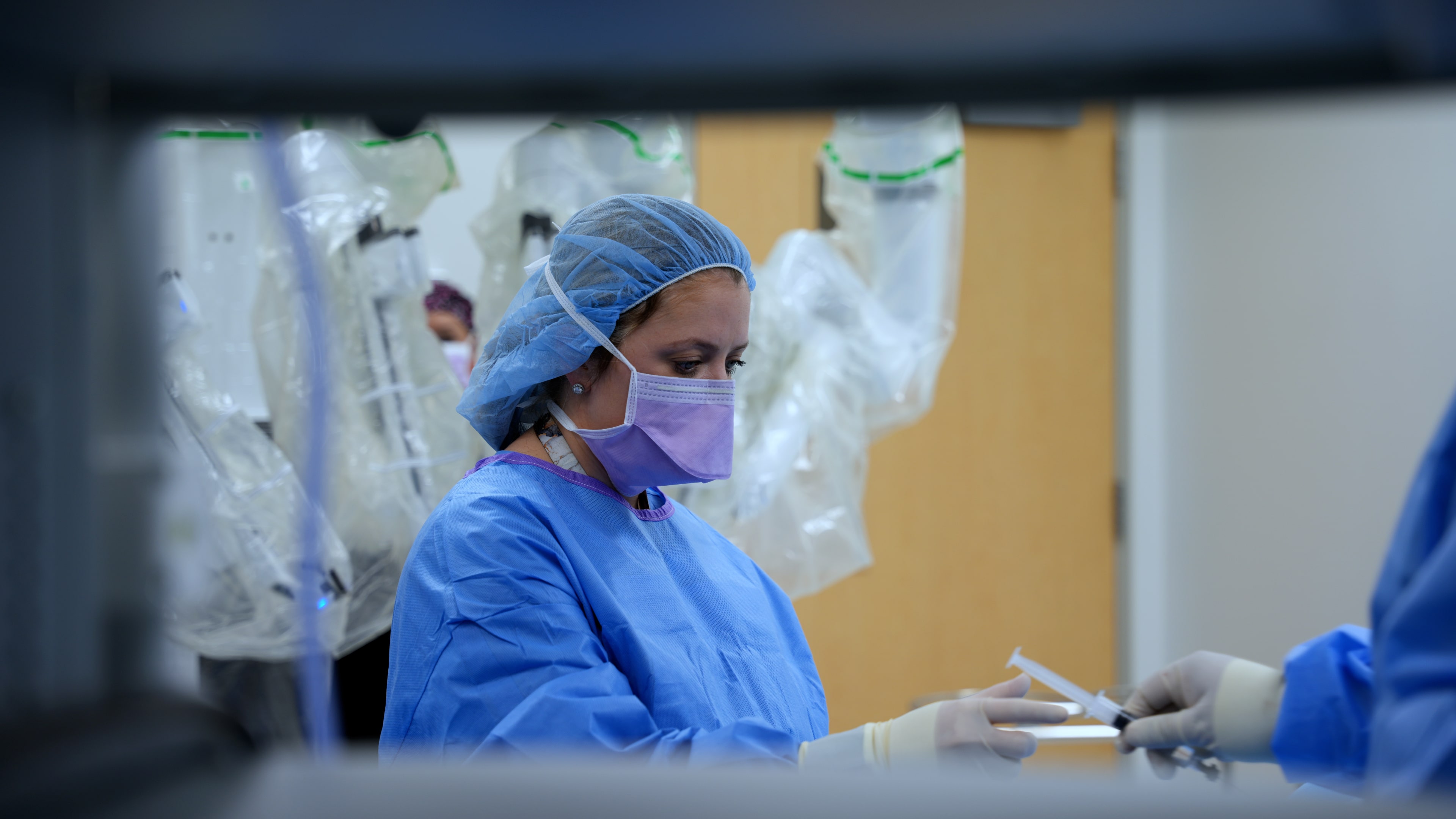 Health care provider wearing a surgical mask, cap and blue gown performs a procedure in an operating room, with medical equipment and surgical instruments visible in the background, illustrating minimally invasive bariatric surgery care