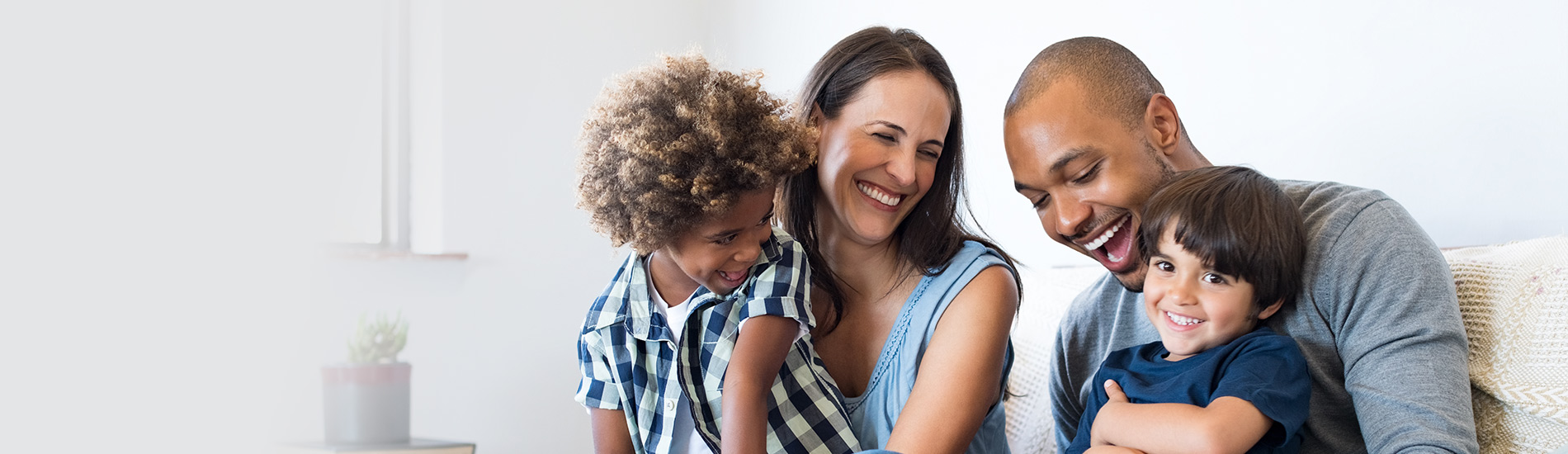 Smiling parents sit on a couch with their two young children, laughing and embracing, showing a happy, healthy family moment at home