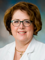 A woman wearing glasses and a white medical coat against a blue-gray studio background is shown in a head-and-shoulders portrait.