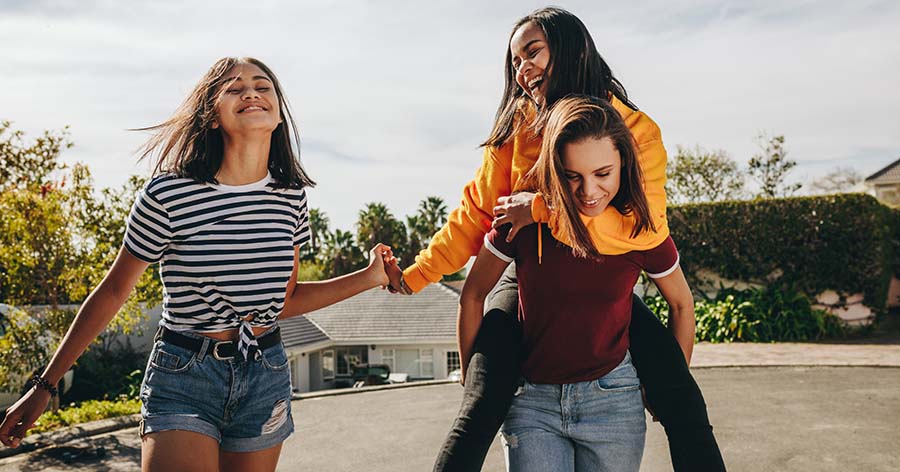 Three youths walk outdoors on a sunny day, with two holding hands and one carrying another on their back, moving along a residential street with houses and trees in the background