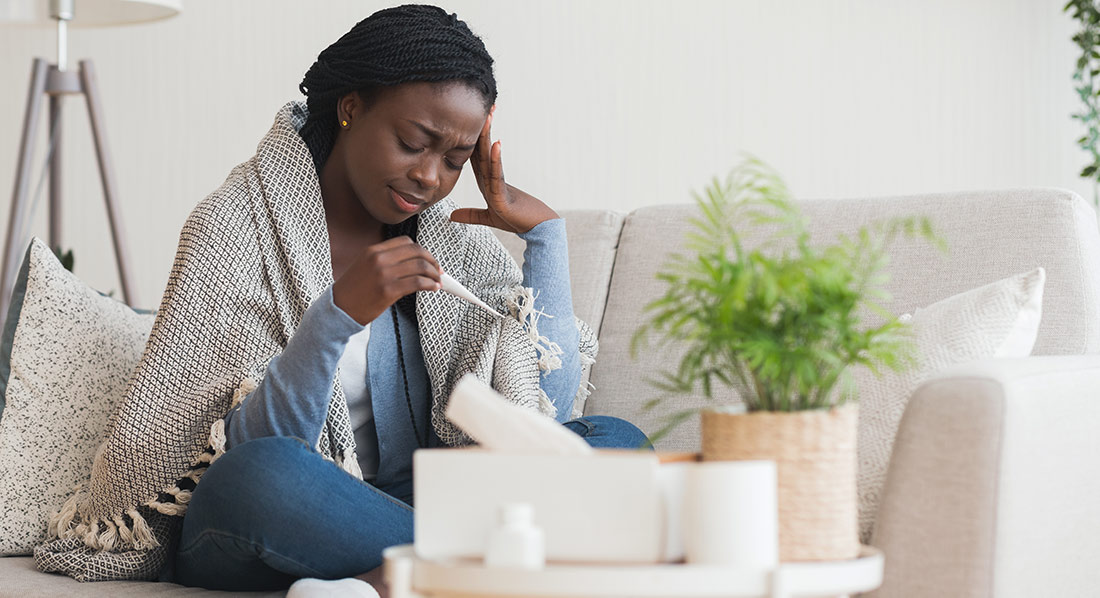A person sits on a couch wrapped in a blanket, holding a thermometer and touching their forehead. A tissue box, cup, and small plant sit on a table nearby in a living room setting.