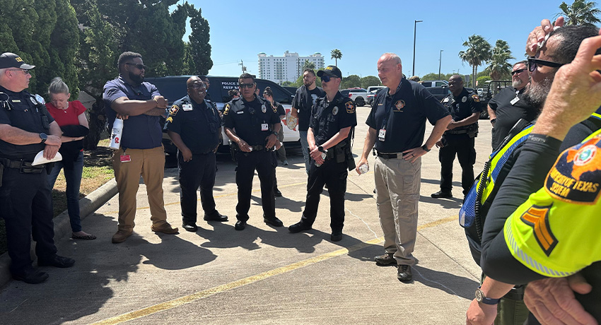 Uniformed law enforcement and public safety personnel stand in a semicircle during a briefing in a parking lot, with vehicles, palm trees, and buildings visible in the background.
